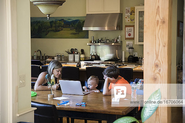 Wide View of Mom am Tisch mit Kleinkind Sohn auf Laptop und Teen Cousin