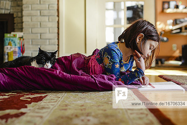 ein Kind  das sich im Schlafsack auf dem Boden zusammenrollt und mit seiner Katze Schularbeiten macht