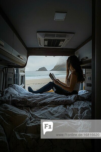 Frau liest auf Bett im Campervan mit Aussicht aus dem Heck vom Campervan auf Strand mit türkisem Wasser  Haukland Strand  Lofoten  Norwegen  Europa