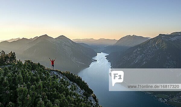 Bergsteiger  junger Mann streckt die Arme in die Luft  Blick über Berglandschaft  Ausblick vom Gipfel des Bärenkopf auf den Achensee  links Seekarspitze und Seebergspitze  Sonnenuntergang in den Bergen  Karwendel  Tirol  Österreich  Europa