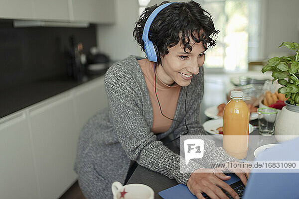 Glückliche Frau mit Kopfhörer arbeiten von zu Hause am Laptop in der Küche