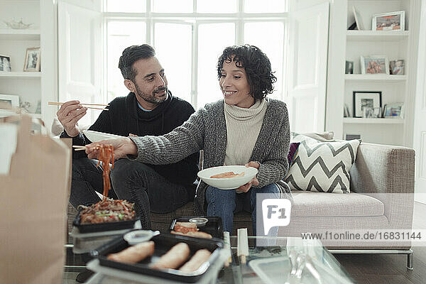 Happy couple enjoying takeout food with chopsticks on living room sofa