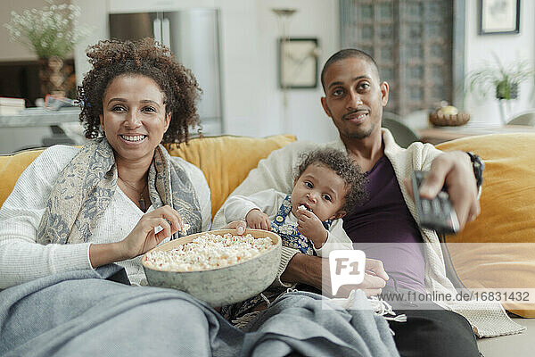 Happy couple with baby daughter watching TV and eating popcorn