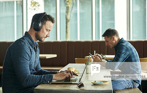 Zwei Männer sitzen in einem Café und arbeiten aus der Ferne mit einem Laptop und einem Smartphone