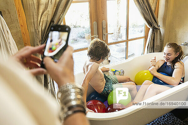 Person  die ein Smartphone-Foto von einem kleinen Jungen und seiner älteren Schwester in der Badewanne mit Wasserballons macht