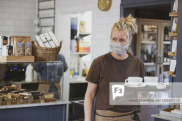 Kellnerin mit Gesichtsmaske arbeitet in einem Café  trägt Tablett mit Kaffeetassen.