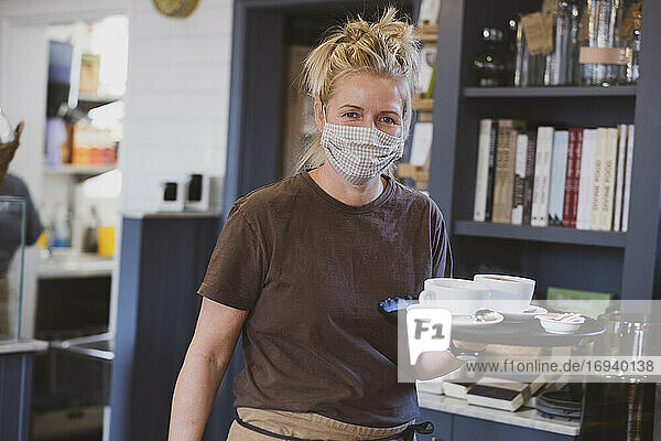 Kellnerin mit Gesichtsmaske bei der Arbeit in einem Café  trägt Tablett mit Kaffeetassen.