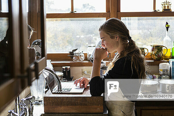 Teenage girl in a kitchen following a baking recipe on a laptop.