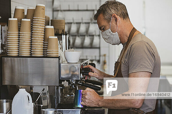 Männlicher Barista mit brauner Schürze und Gesichtsmaske  der in einem Café arbeitet und Espresso zubereitet.