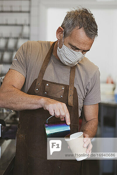 Männlicher Barista mit brauner Schürze und Gesichtsmaske arbeitet in einem Café und schenkt Kaffee ein.