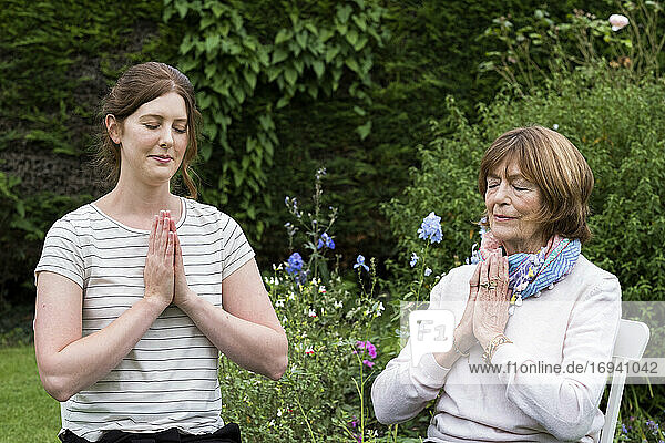 Frau und Therapeutin in einem Garten sitzend  Hände zusammen und Augen geschlossen.