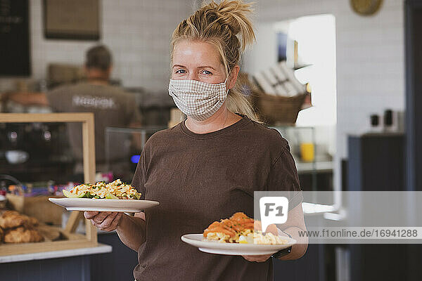 Kellnerin mit Gesichtsmaske bei der Arbeit in einem Café  trägt Teller mit Essen.
