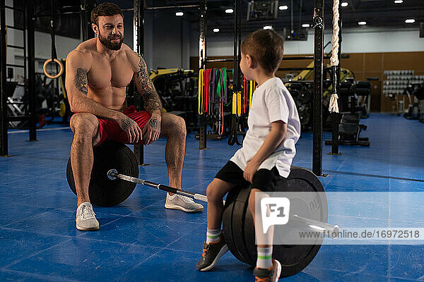 Vater und Sohn sitzen während einer Pause im Fitnessstudio auf einer Langhantel