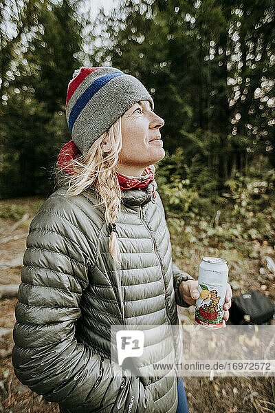 Smiling happy blonde woman in gray jacket and winter hat holding beer.