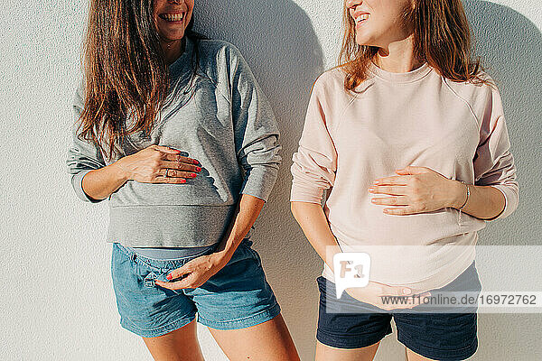 Zwei junge schwangere Frauen halten ihren Bauch und lächeln sich gegenseitig an