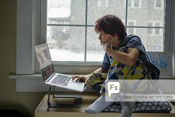 Ein ernster Junge sitzt auf einer Bank am verschneiten Fenster und arbeitet am Computer