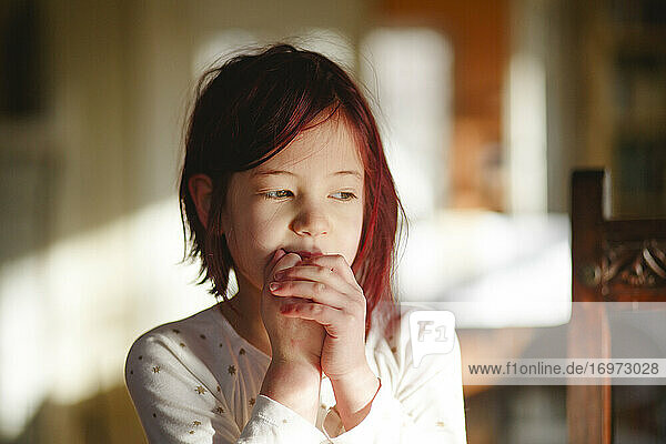 A shy little girl with dyed red hair sits alone with hands clasped