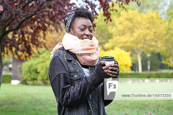 Schöne afrikanische Frau  die einen Schal trägt und eine Tasse Kaffee in einem Park hält. Herbst  Winter und kalte Konzepte.