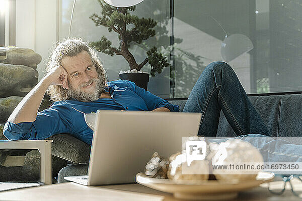 Älterer Mann mit Bart telefoniert und trinkt Kaffee  sitzt auf einem Sofa vor seinem Laptop und schaut aus dem Fenster. Geschäftskonzept
