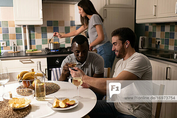 Fröhliche Männer benutzen ihr Smartphone  während die Frau das Abendessen kocht