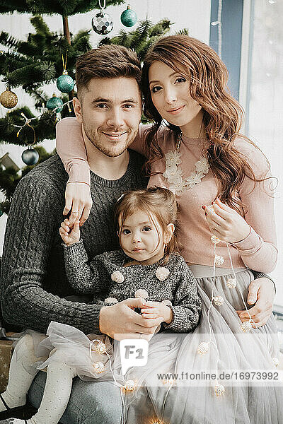 Young family with a daughter in festive outfits with a garland near the Christmas tree on New Years Eve