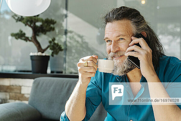 Älterer Mann trinkt einen Kaffee und telefoniert auf dem Sofa in seinem Wohnzimmer. Business-Konzept