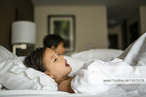 Ein kleines Mädchen liegt in einem Hotelzimmerbett und singt Bruder im Hintergrund