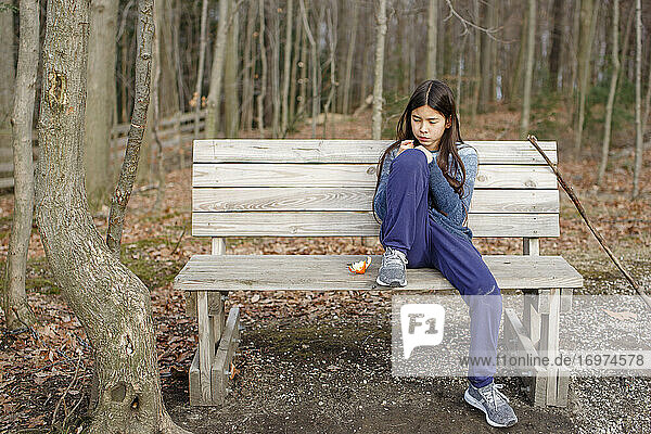Ein kleines Mädchen sitzt allein auf einer Bank im Wald und isst eine Orange im Herbst