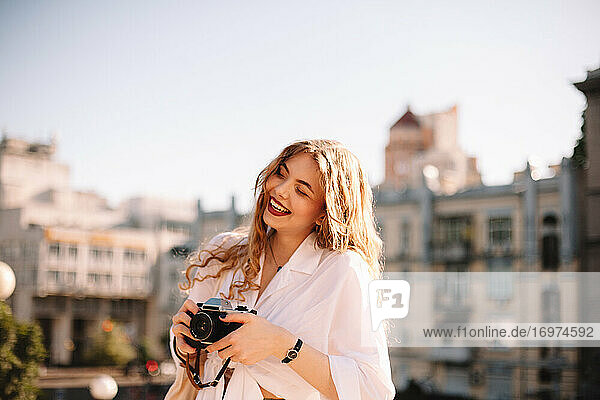 Glückliche junge Frau  die eine Kamera hält  während sie im Sommer in der Stadt steht