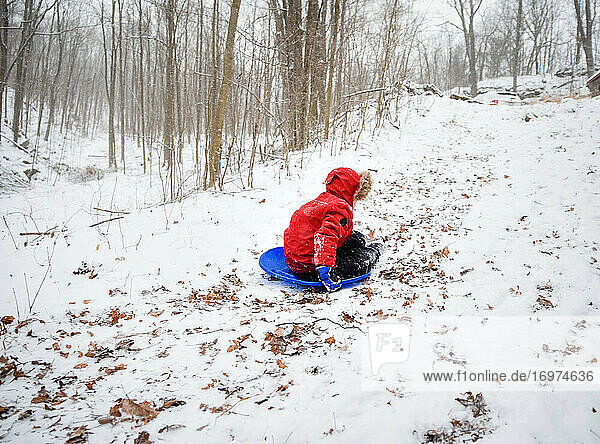Boy in red coat sledding down hill in the woods on a snowy winter day.