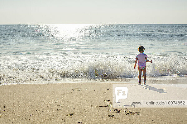 A little girl stands at the edge of the shore with oncoming wave