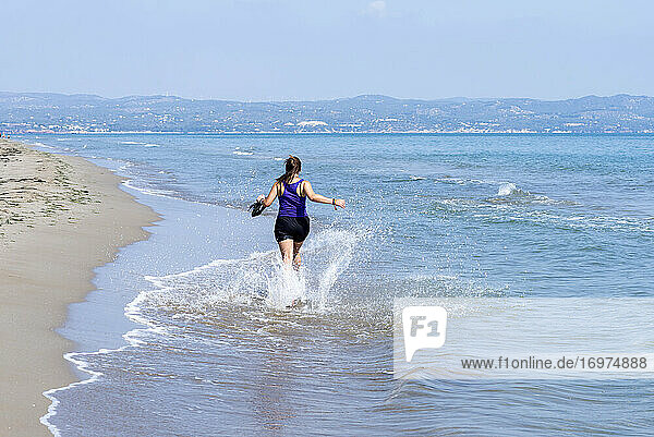 Rückenansicht einer Frau in Shorts  die am Meeresufer läuft und Wasser spritzt