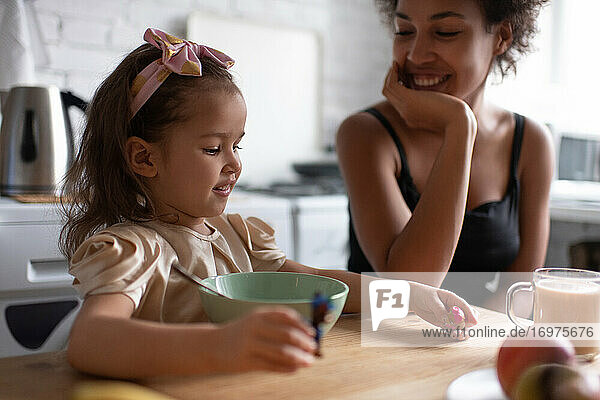 Fröhliches ethnisches Mädchen spielt beim Frühstück mit seiner Mutter