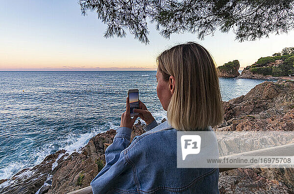 Junges blondes Mädchen beim Fotografieren des Sonnenuntergangs an der Küste