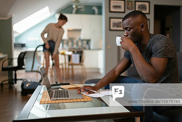 Schwarzer Freiberufler mit Laptop  während seine Freundin das Wohnzimmer aufräumt