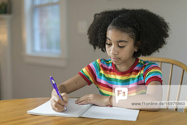 Zehnjähriges gemischtrassiges Mädchen schreibt am Tisch in ein Notizbuch