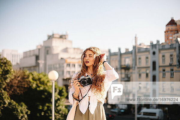 Nachdenkliche junge Frau hält eine Kamera  während sie im Sommer in der Stadt steht