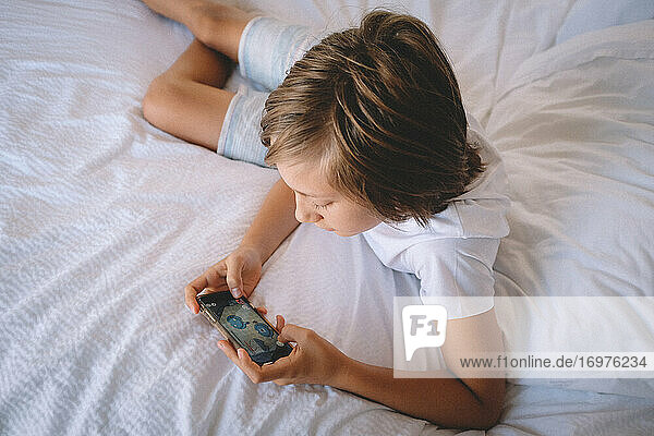 Boy in White Checks spielt auf seinem Handy von einem Hotelzimmerbett aus.