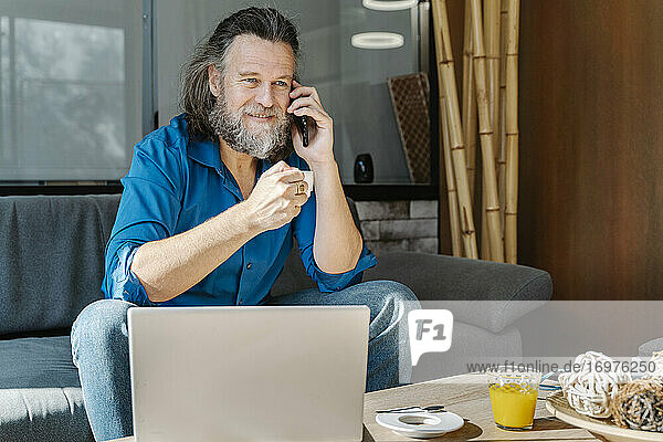 Mature man with beard talking on the phone and drinking coffee sitting