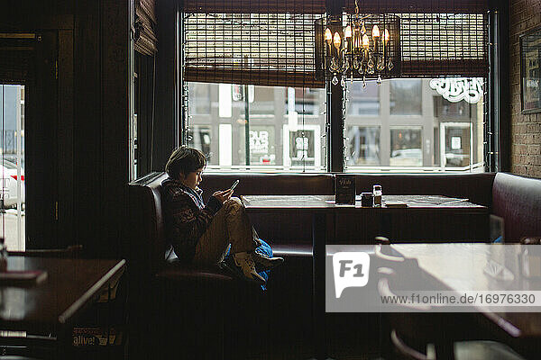 Ein kleiner Junge sitzt an einem Restauranttisch am großen Fenster und schaut auf sein Handy.