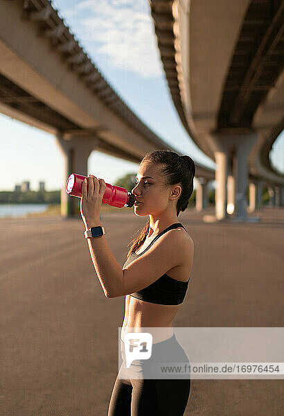 Schlanke Sportlerin trinkt Wasser am Ufer unter einer Brücke