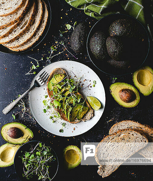 Avocado toast on a plate with ingredients around it on black counter.