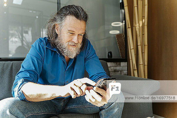 Älterer Mann mit Bart sitzt auf einem Sofa und schaut auf sein Smartphone im Wohnzimmer seines Hauses. Business-Konzept