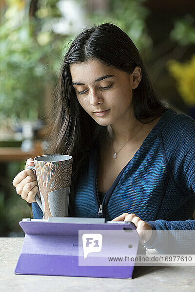 Hübsches Teenager-Mädchen arbeitet am iPad und hält eine Kaffeetasse