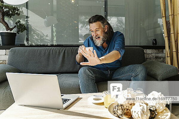 Älterer Mann mit Bart lächelt und arbeitet mit seinem Laptop auf einem Sofa zu Hause. Business-Konzept