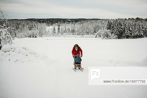 Mother pushing child on sleigh in snow in woods on cold winter day