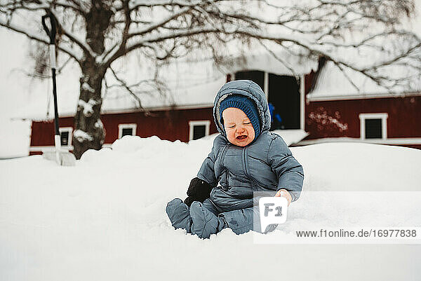 Baby crying sitting on snow in Norway with red barn behind
