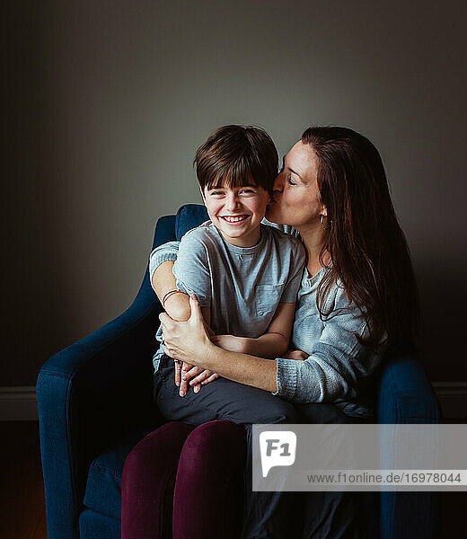 Eine Frau umarmt und küsst ihren Sohn  der in ihrem Schoß auf einem Stuhl sitzt.