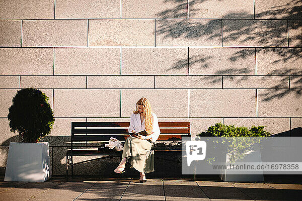 Young woman writing in diary sitting on bench by wall in summer city