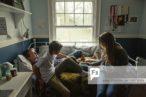 drei Geschwister sitzen auf einem Bett und kuscheln mit einem süßen Familienhund im Schlafzimmer